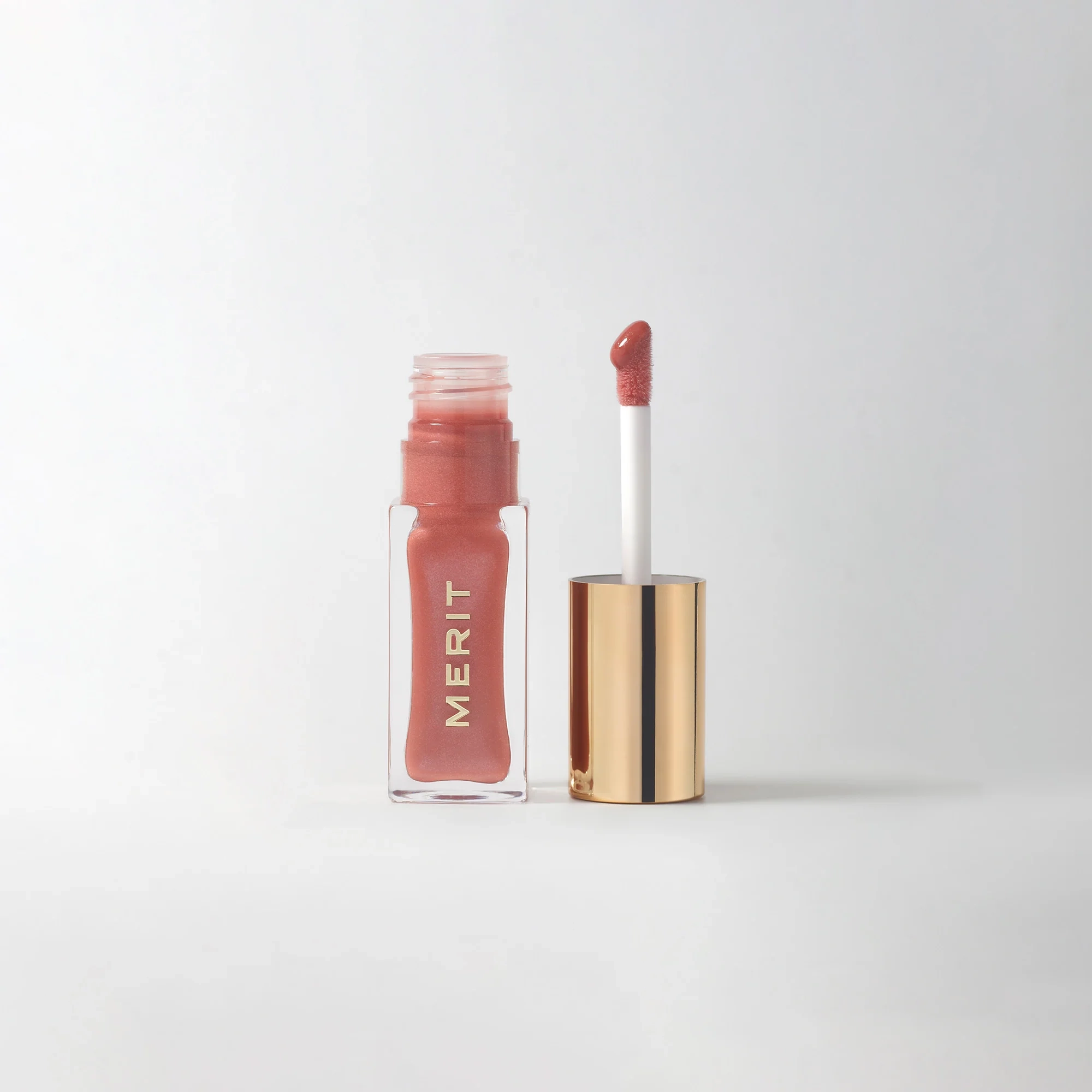 A bottle of MERIT lip oil in a pearlescent pink shade next to its applicator